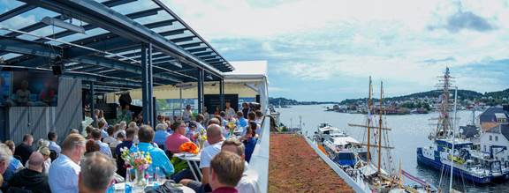 Audience attending ENRX rooftop event during Arendalsuka 2024 in Arendal, Norway, discussing sustainable energy transfer with harbor view and tall ships in background.